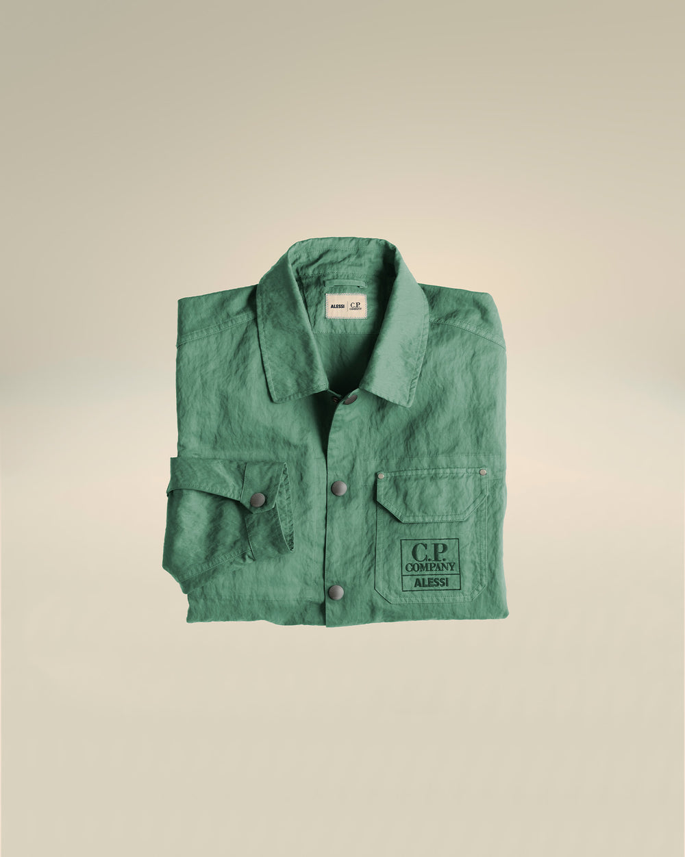 Green utility jacket by Alessi with a crinkled texture, featuring a C.P. Company ALESSI logo on the chest pocket, folded flat.