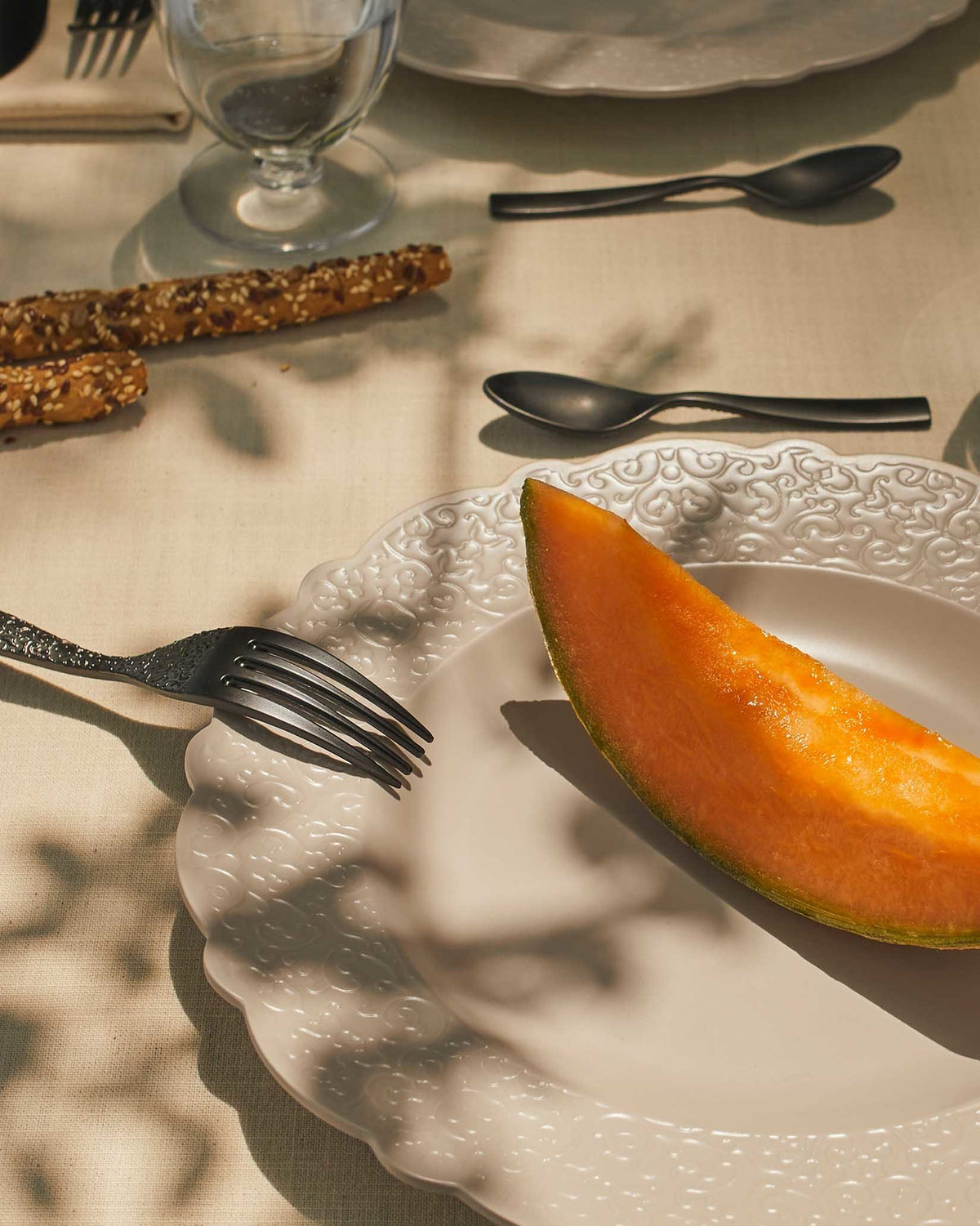 Ornate white plate by Alessi with scalloped edge, embossed scrollwork, black fork, and a cantaloupe slice.